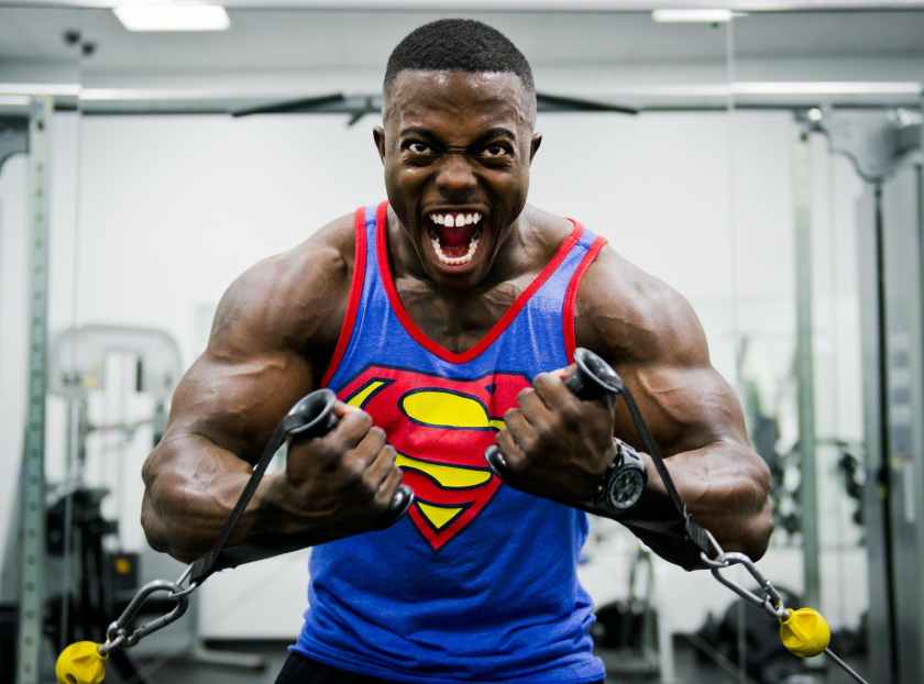 Veiny body builder with his mouth  open and wearing a superman tank is intensely doing a low cable chest fly.